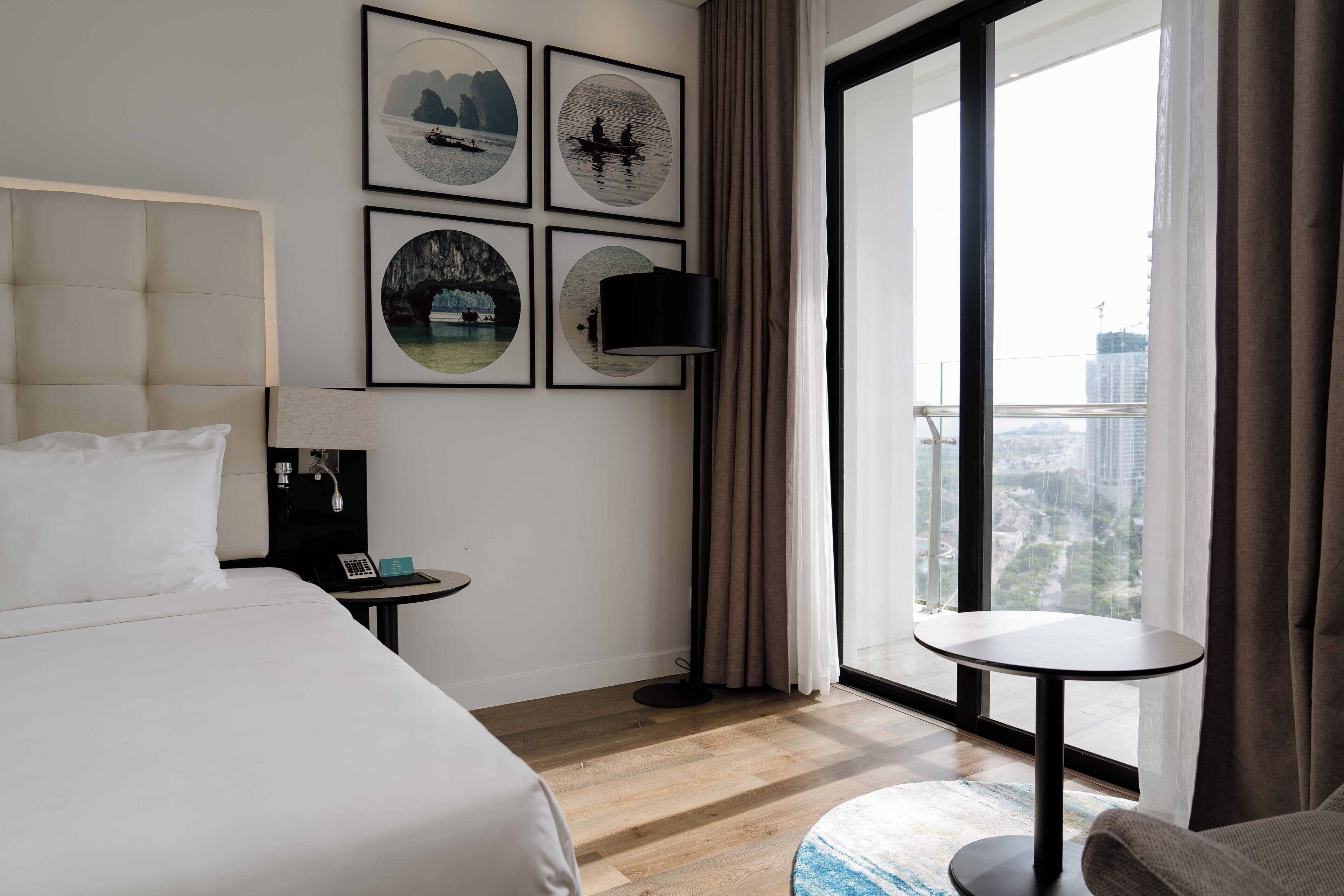 A hotel room with a bed, a bedside table with a phone, a small round table, a wall-mounted TV, and framed pictures.
