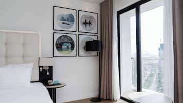 A modern hotel room with a large bed, a bedside table with a lamp, a small table, and a large window with a view of the city.