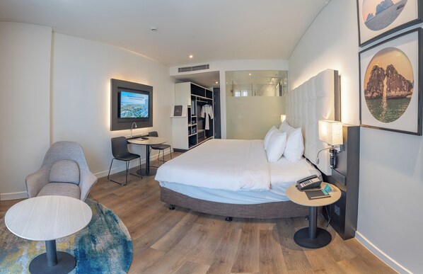 A modern hotel room with a large bed, a desk, a chair, a small table, and a TV.