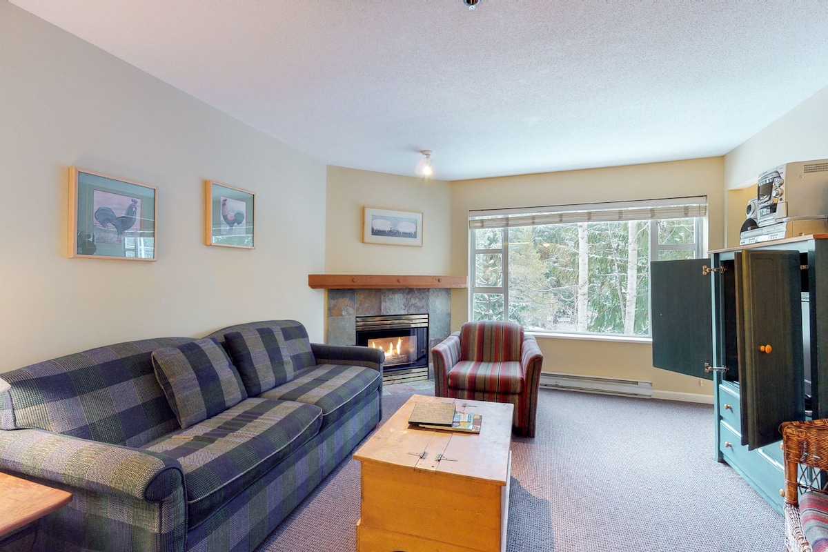 A living room with a sofa, armchair, coffee table, fireplace, and a large window with a view of trees.