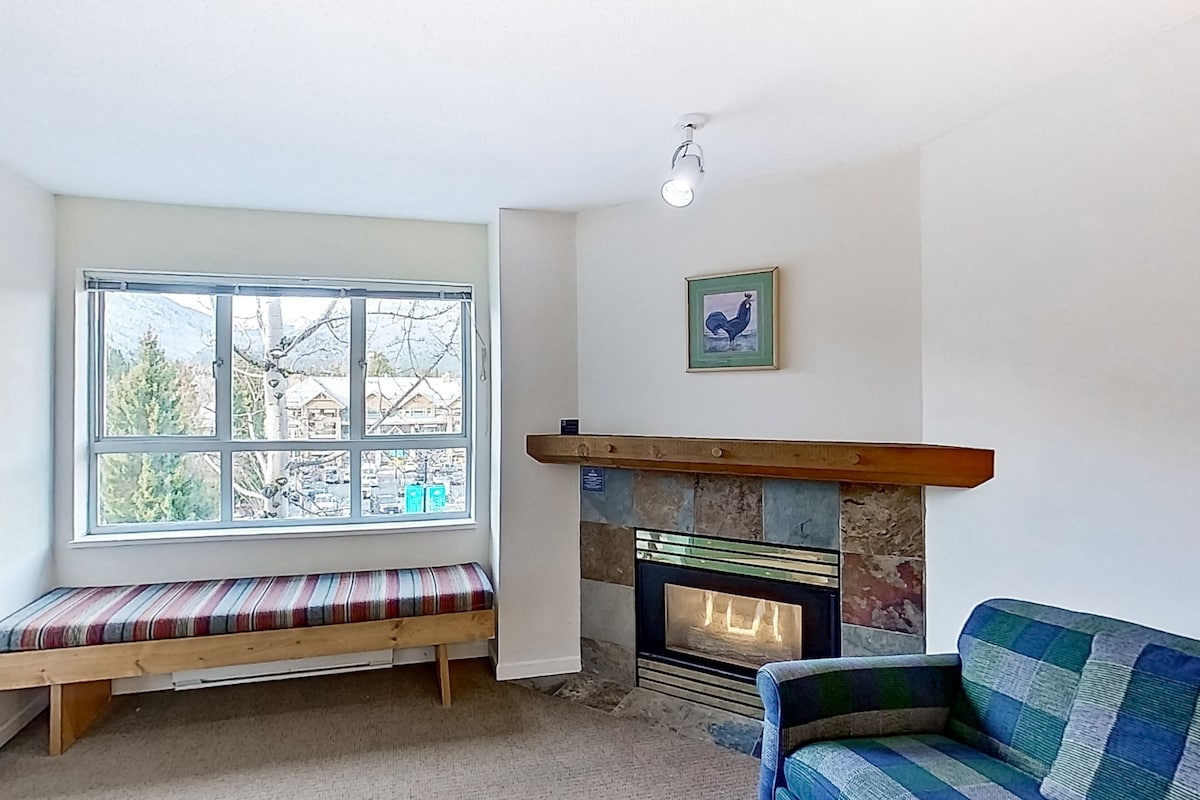 A cozy living room with a bench, a fireplace, and a large window offering a view of the outdoors.