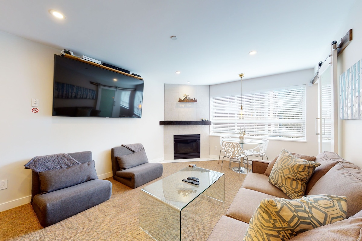 A modern living room with a glass coffee table, sofas, a fireplace, and a dining area.
