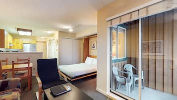 Une chambre d’hôtel compacte comprenant un lit, un petit bureau, une chaise, un canapé, une kitchenette et une porte-fenêtre coulissante donnant sur un balcon équipé de chaises.