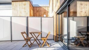 A modern outdoor patio with a wooden table and chairs, a glass partition, and a view of the adjacent building. - Apartment Comfortable in the centre of Pau: parking, AC... (Pau)
