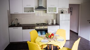 A modern kitchen with white cabinets, a gas stove, and a refrigerator. There is a dining table with yellow chairs and a fruit bowl.
