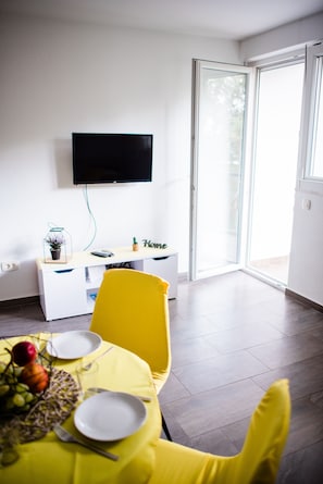 A modern room with a flat-screen TV mounted on the wall, a sliding glass door leading to an outdoor area, a yellow chair, a dining table with plates and cutlery, and a small potted plant on a white console table.