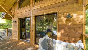 Eine Holzhütte mit einer Terrasse, großen Glasfenstern und einem gemütlichen Innenraum mit einer Couch und Kissen.