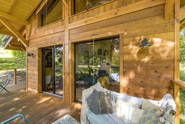 A wooden cabin with a deck, large glass doors, and a cozy interior with a sofa and cushions.
