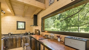 Ein Holzblockhaus-Interieur mit Küchenzeile, Barhocker, einem großen Fenster mit Blick auf einen Wald und einem Vintage-Marshall-Lautsprecher.