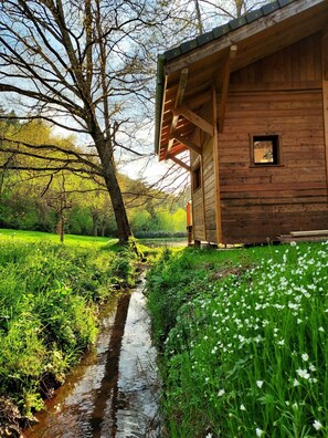 A wooden cabin with a small window, surrounded by lush greenery and a stream.