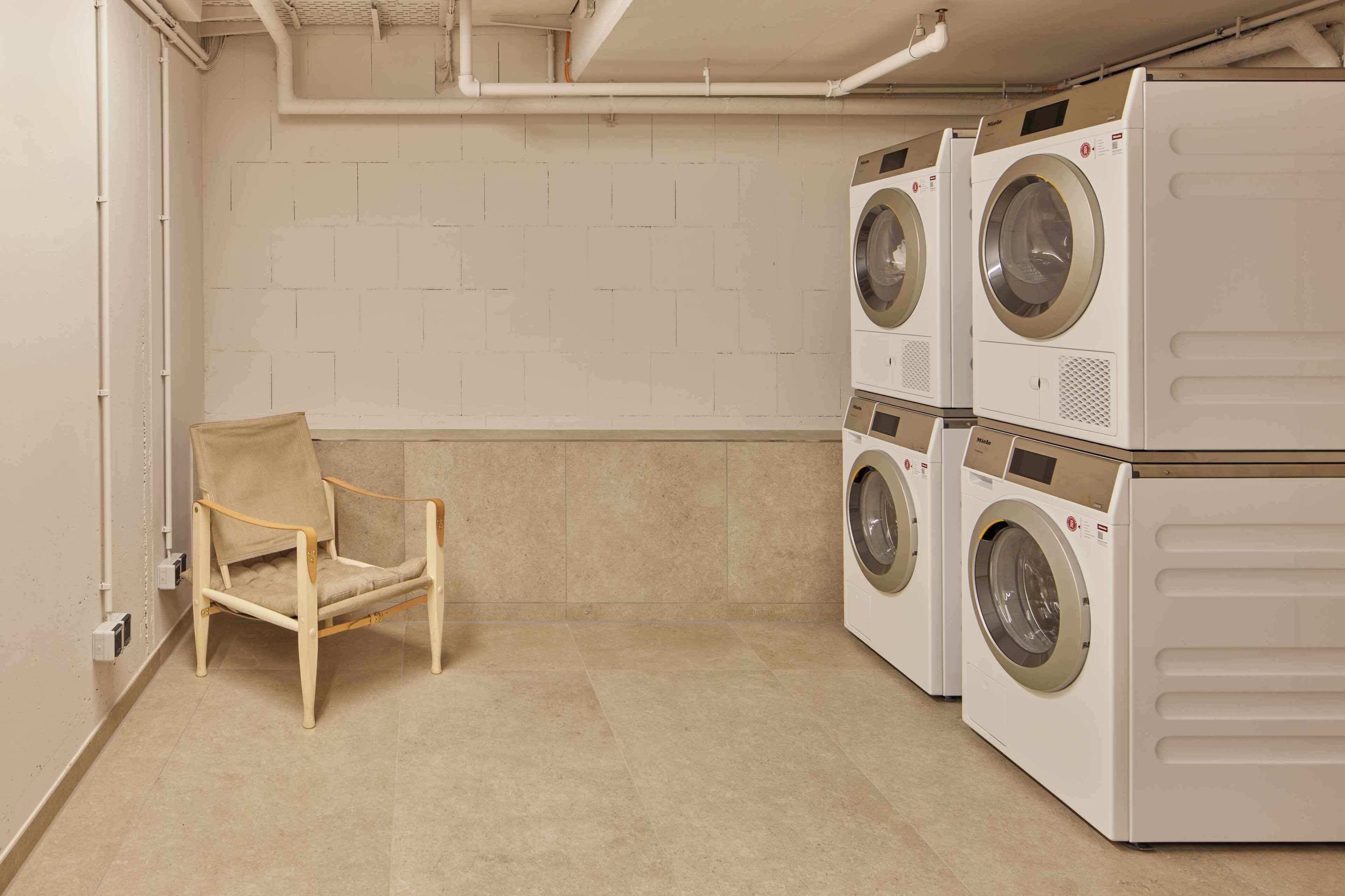 A laundry room with a chair, two stacked washing machines, and a white tiled wall.
