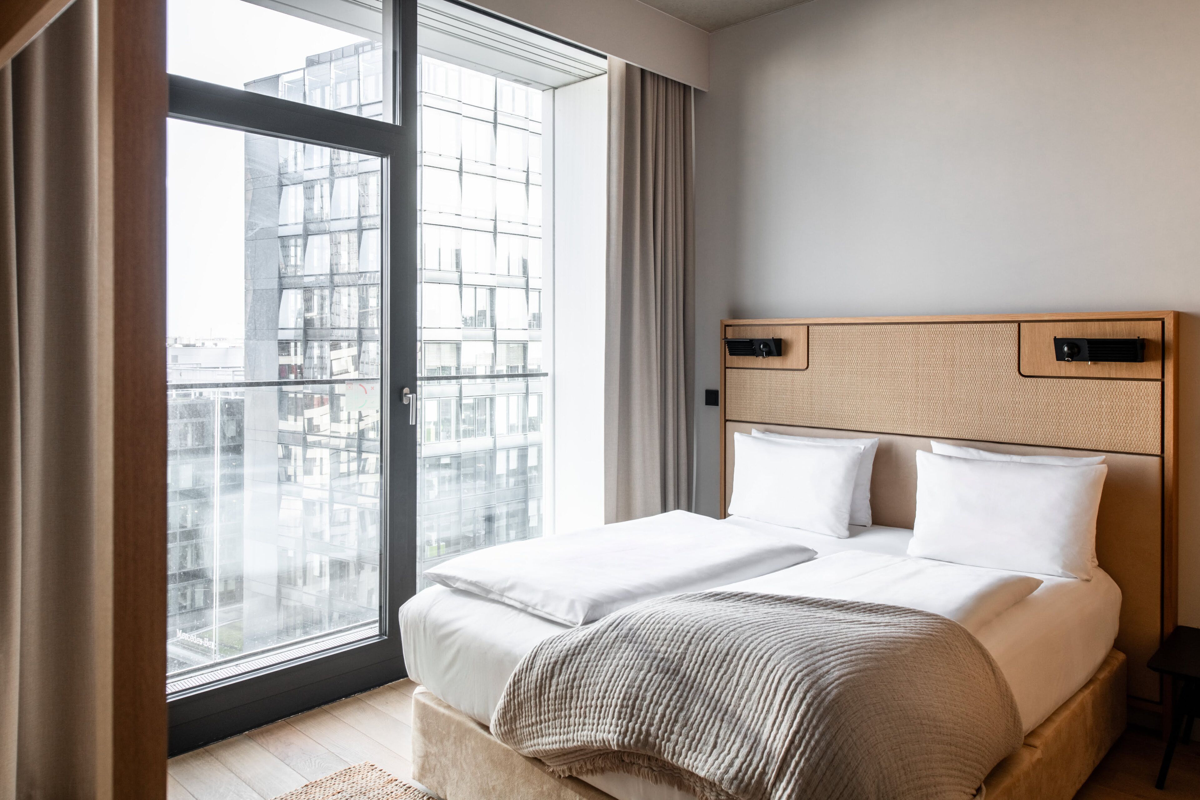 Una habitación de hotel moderna con un ventanal grande, una cama con ropa blanca, cabecera de madera y vistas a un paisaje urbano.