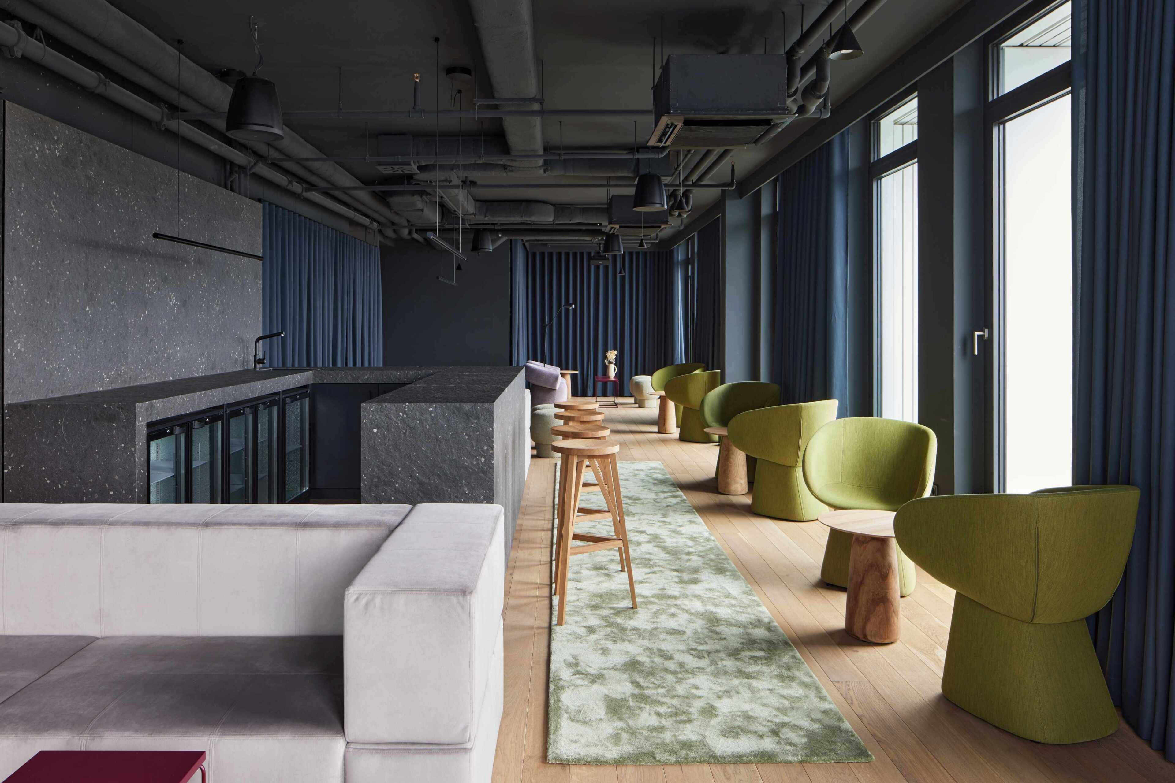 A modern lounge area with a sofa, green chairs, a wooden stool, and a patterned rug.