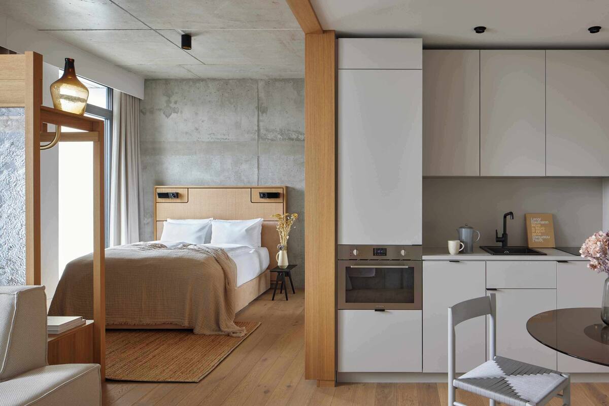 A modern hotel room with a bed, a small table with a chair, a kitchenette, and a large window.
