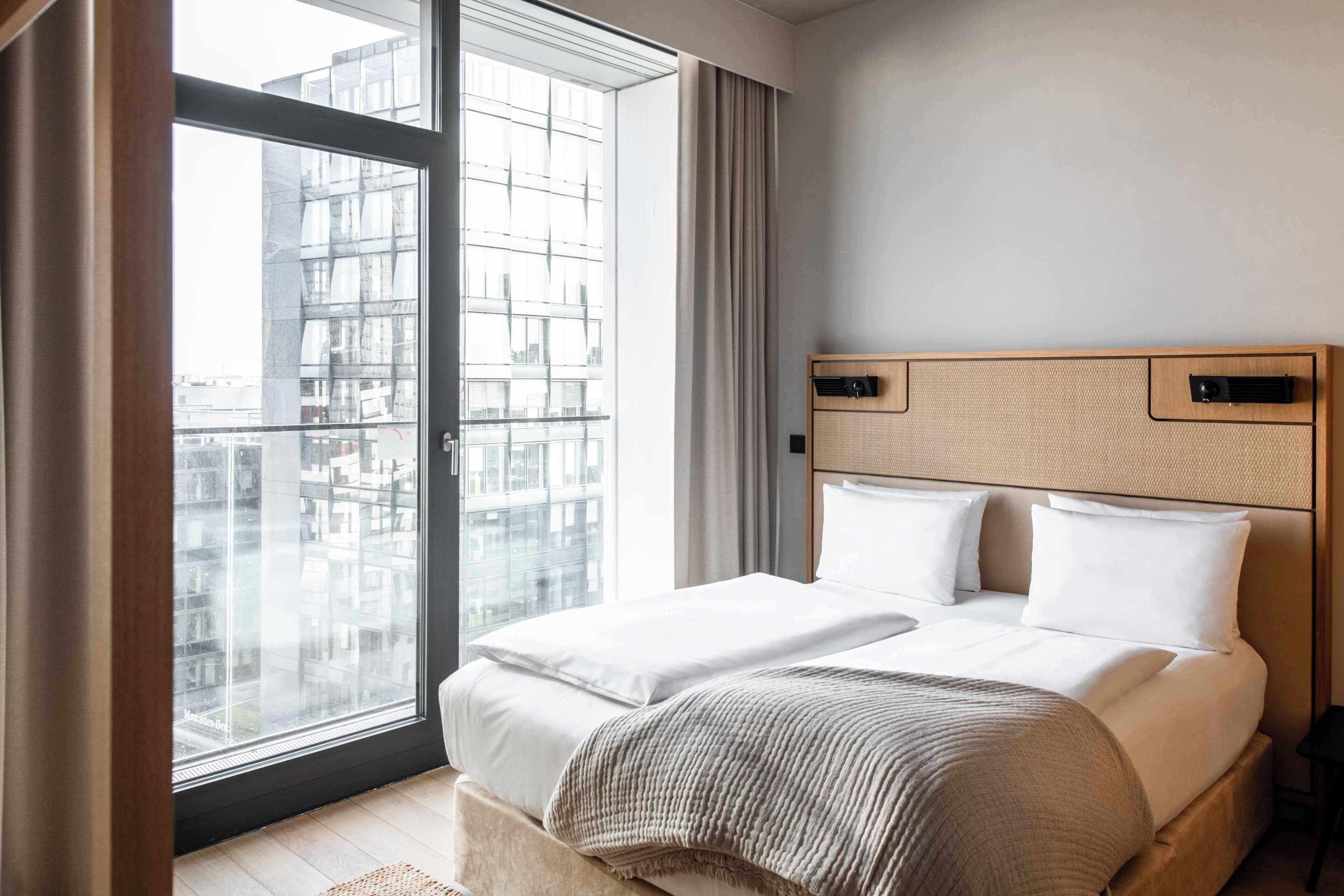 A modern hotel room with a large window, a bed with white linens, a wooden headboard, and a view of the cityscape.