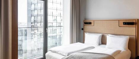 A modern hotel room with a large window, a bed with white linens, a wooden headboard, and a view of the cityscape.