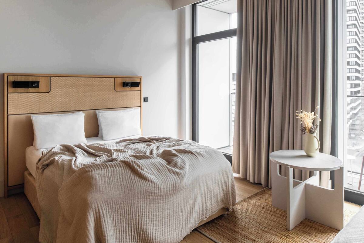 A modern hotel room with a large bed, a small round table, and a view of the city through large windows.