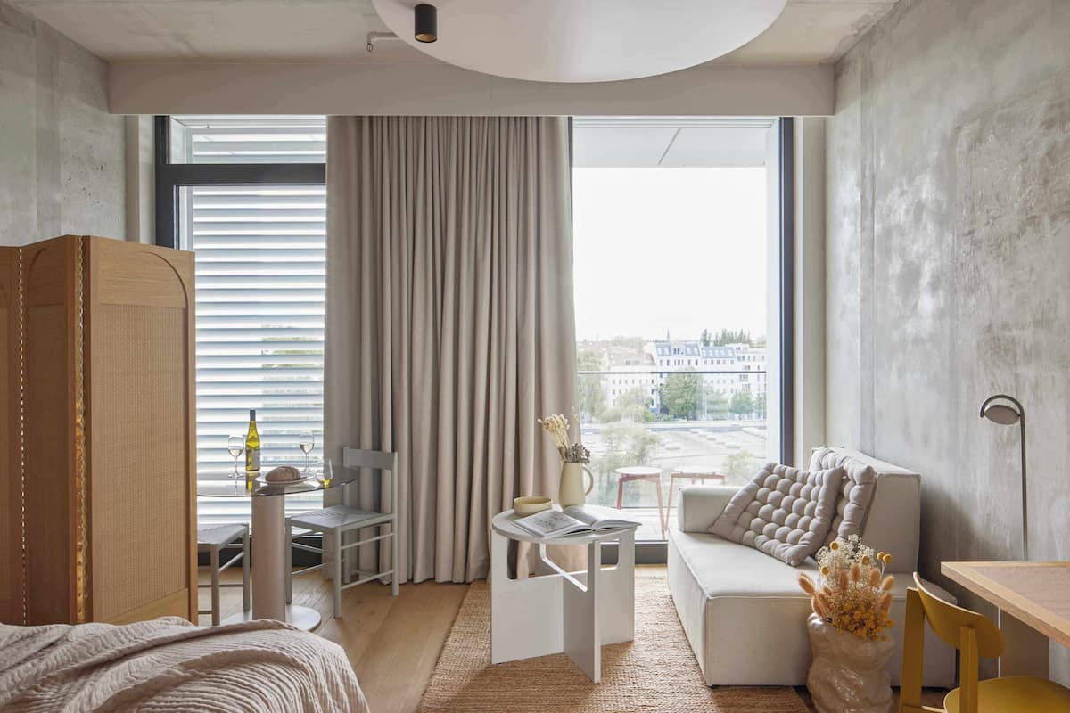 A modern hotel room with a large window, a sofa, a dining table, and a small balcony.