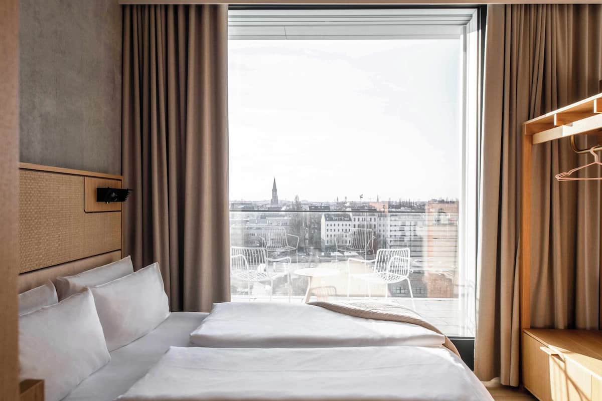 A hotel room with a large window, a bed, a bedside table, and a view of the cityscape.