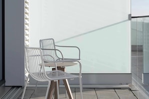 A balcony with two white metal chairs and a table, a glass railing, and a white wall. - Locke at East Side Gallery (Berlin)