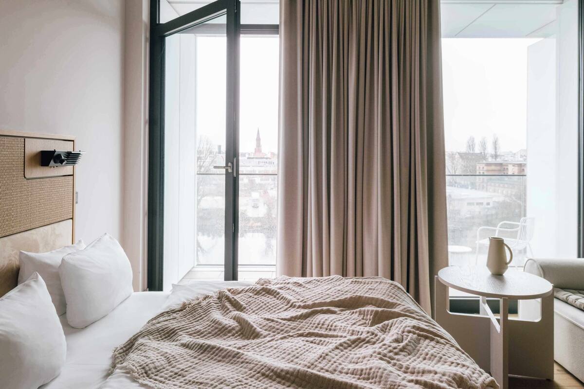 A hotel room with a bed, a small table, a chair, and a view of the city through a large window.