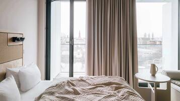 Habitación de hotel con cama, una mesita, una silla y vista a la ciudad a través de un ventanal grande.