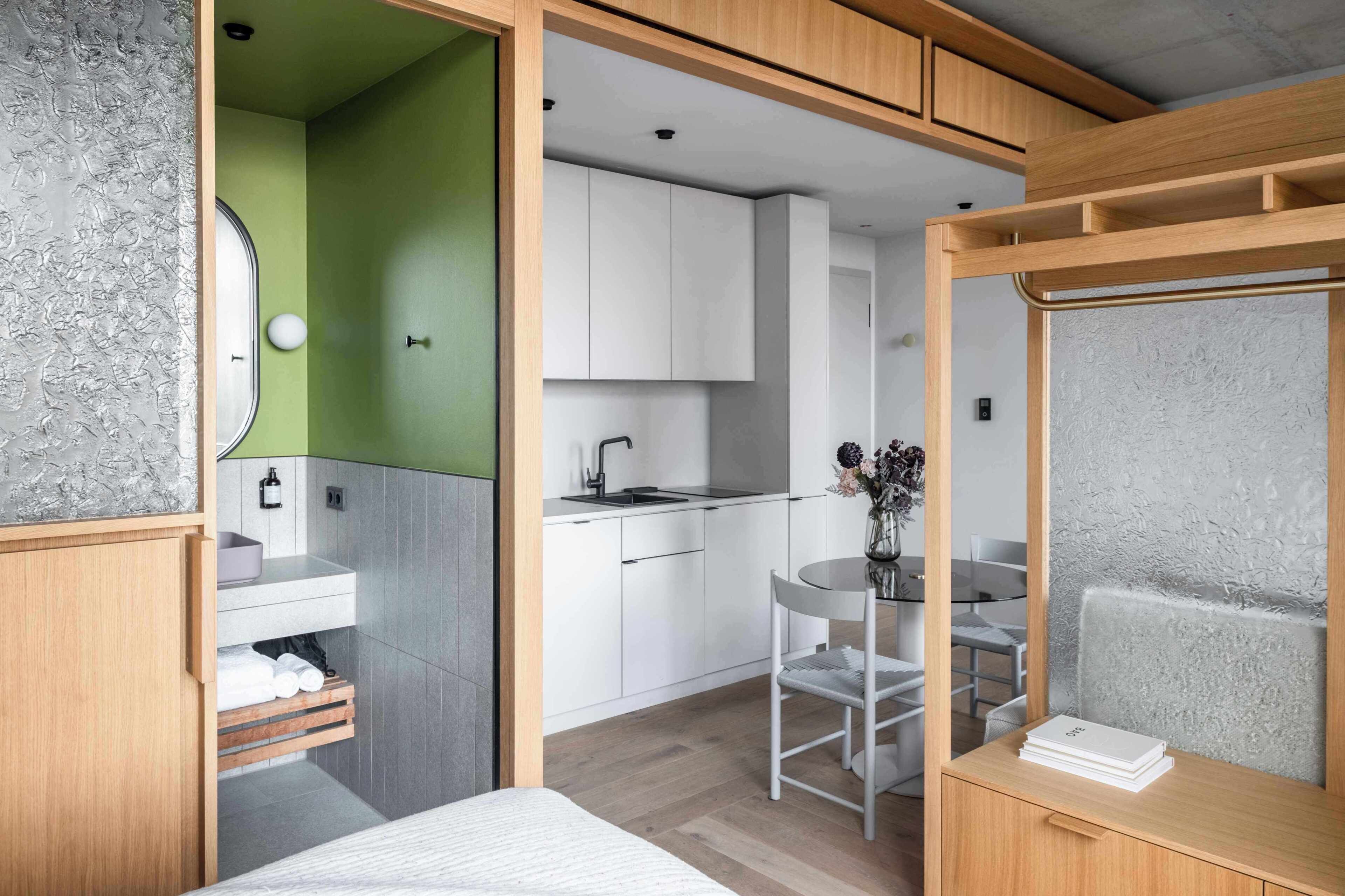 A modern room with a bed, a small kitchenette, a bathroom with a green wall, and a seating area with a table and chair.