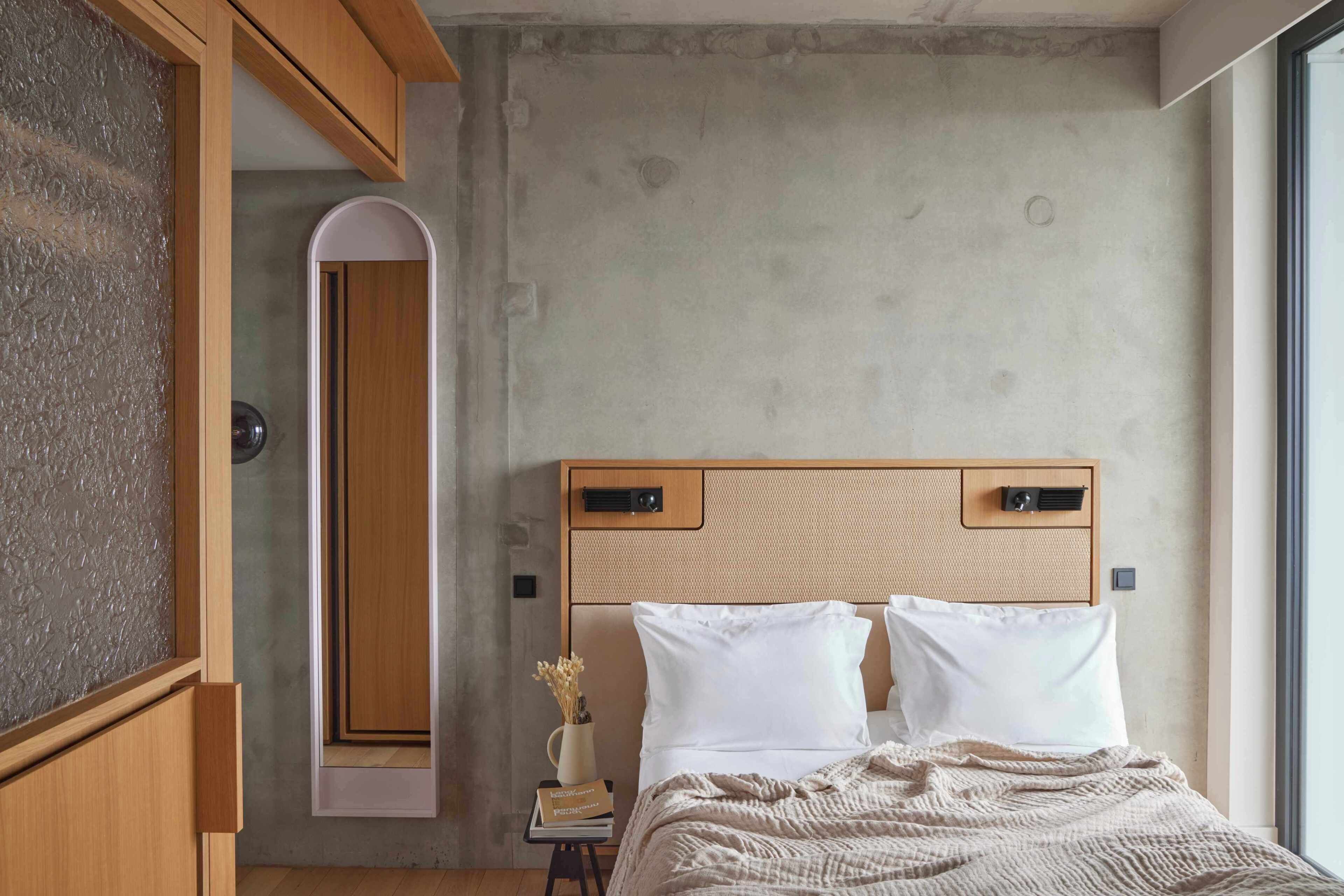 A bedroom with a wooden headboard, a bedside table with a book and a vase, and a large window.