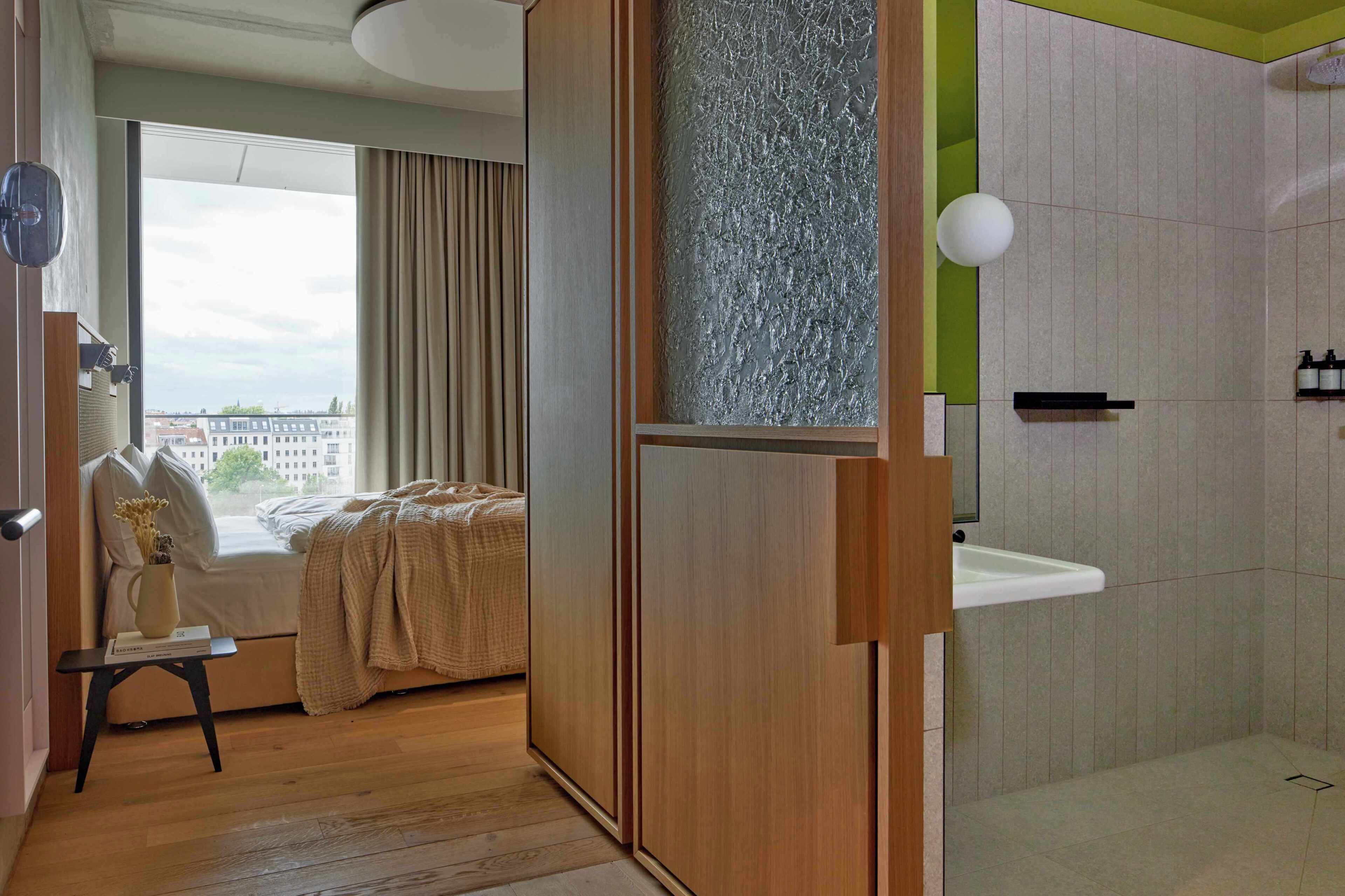 A room with a bed, a window with curtains, a wooden door, a bathroom with a sink, and a city view.