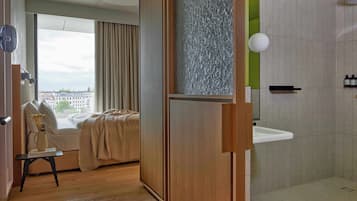 A room with a bed, a window with curtains, a wooden door, a bathroom with a sink, and a city view.