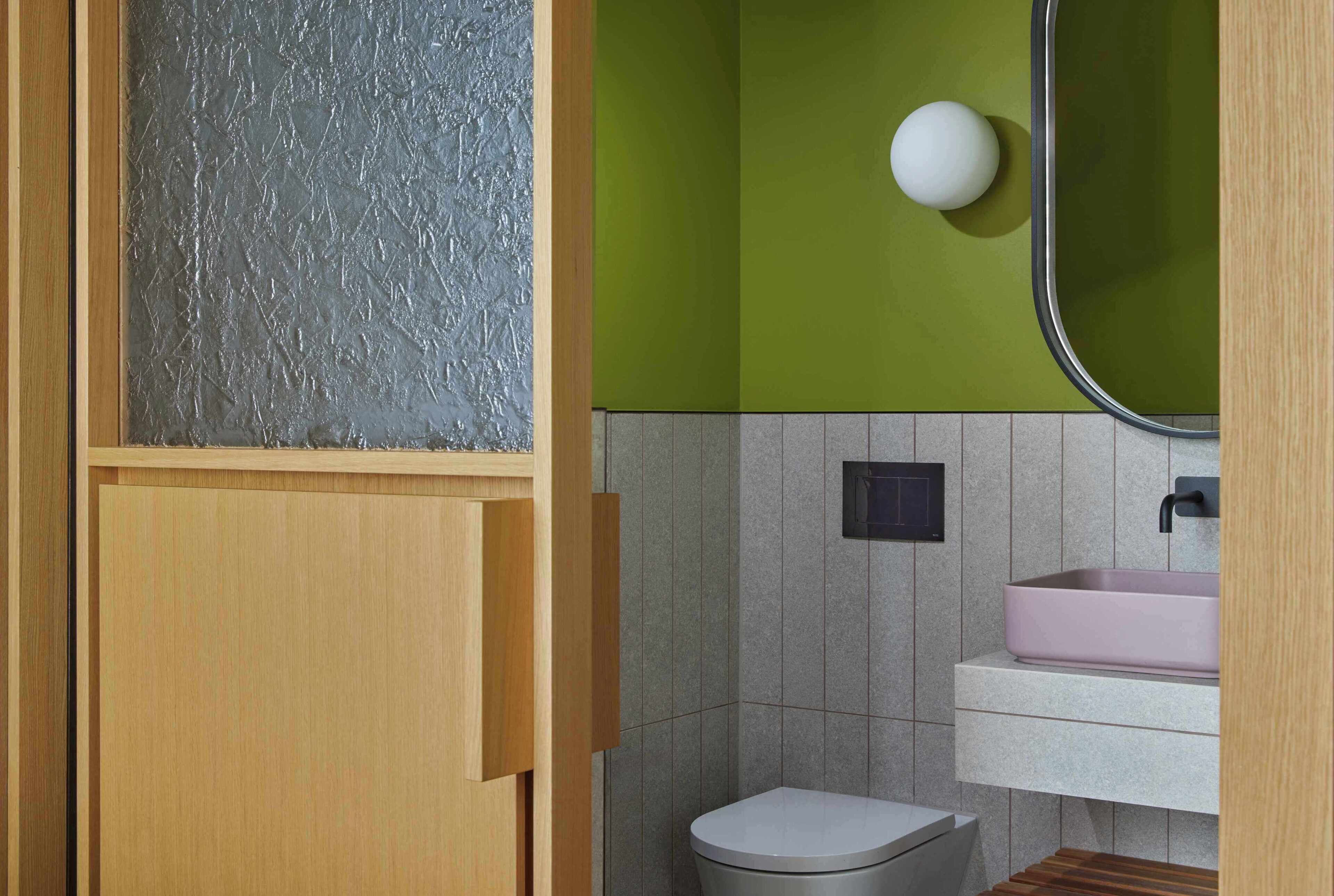 Ein Badezimmer mit einem Holzkosmetikschrank, einer weißen Toilette, einem pinken Waschbecken und einer grünen Wand.