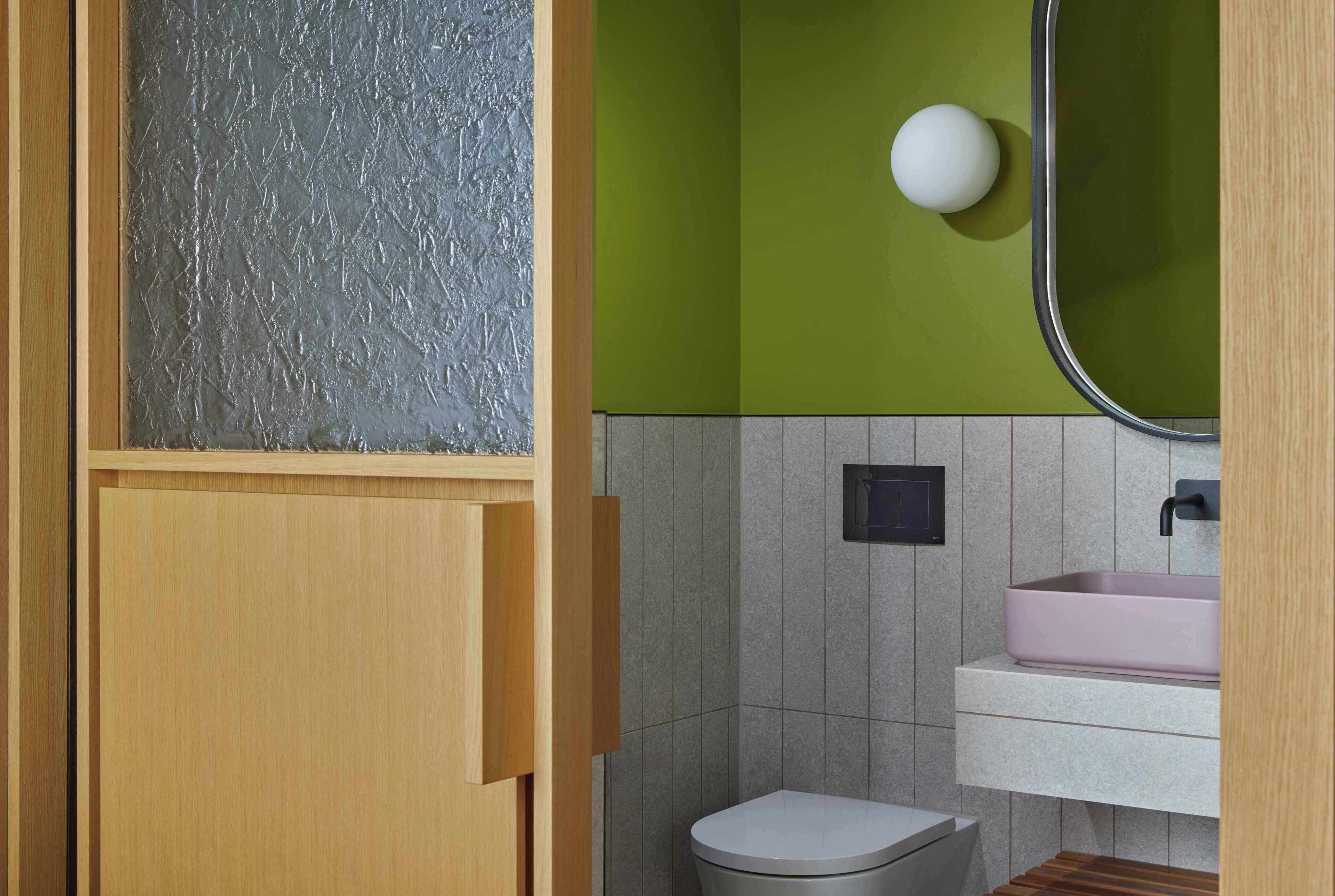 A bathroom with a wooden door, a green wall, a white toilet, a pink sink, and a mirror.