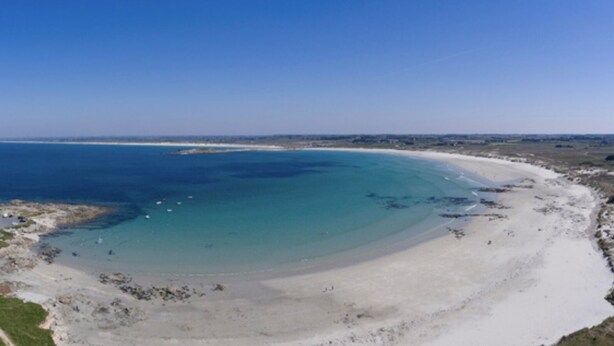 A coastal landscape with a sandy beach, a clear blue sea, and a road leading to a white building.