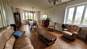 A spacious living room with a leather sofa, a coffee table, and leather armchairs. There is a dining table with chairs, a chandelier, and large windows providing natural light.