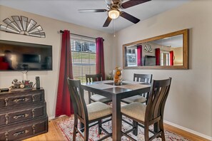 A dining area with a wooden table, four chairs, a ceiling fan, a large mirror, and a TV on a wall.