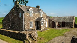 Une maison en pierre à l’architecture traditionnelle, dotée d’une grande fenêtre et d’une porte, située dans un paysage rural avec un chemin de gravier et une zone herbeuse.