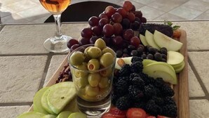 Quarto com cama, mesa com tábua de frutas e uma taça de vinho, e vista para o oceano.