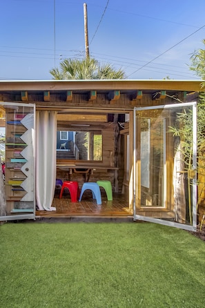 A wooden structure with a sliding door, a small seating area with colorful stools, and a view of a house through the door.