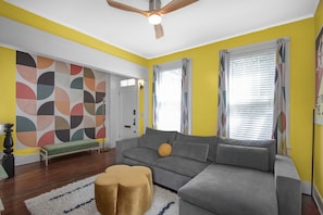 A living room with a grey sofa, a wooden floor, a ceiling fan, and a colorful wall design.