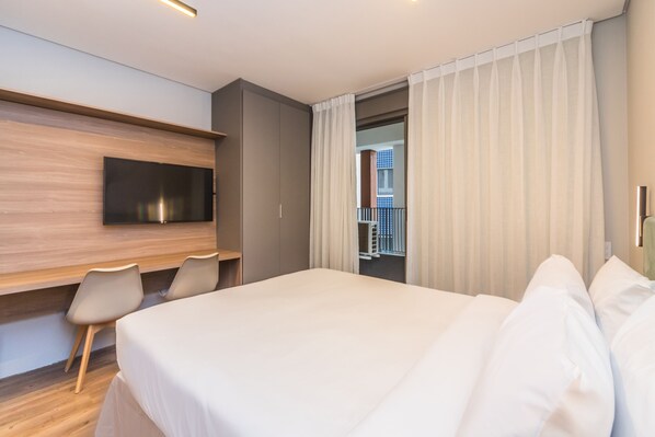 Une chambre d’hôtel moderne dotée d’un grand lit, d’un téléviseur fixé au mur, d’un bureau avec des chaises et d’un balcon avec une rambarde.