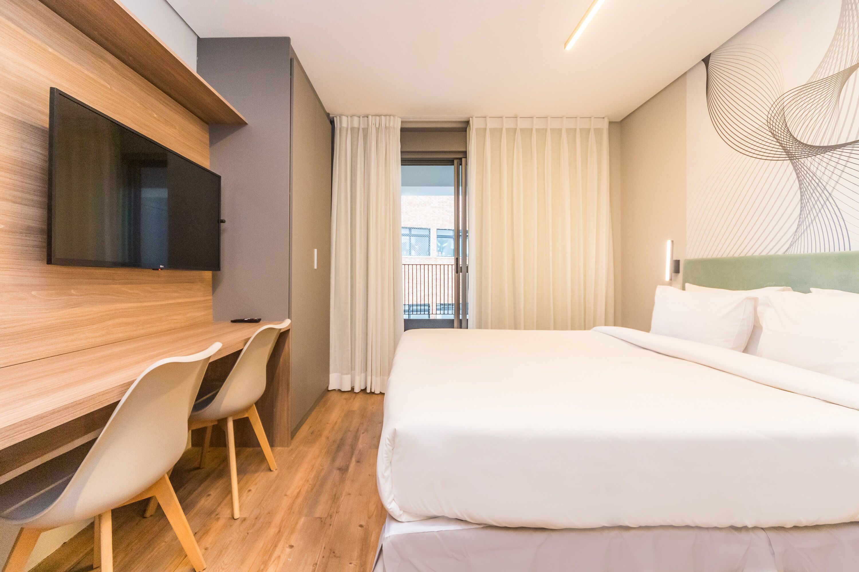 Una habitación de hotel moderna con un escritorio de madera, dos sillas, un televisor de pantalla plana, una cama grande y vistas al exterior a través de una puerta de balcón.