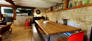 Uma área de jantar rústica com mesa de madeira, bancos, paredes de pedra e uma cozinha ao fundo.