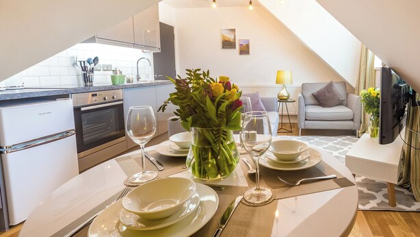 Een eethoek met een gedekte tafel voor twee personen, een bloevas en een keuken op de achtergrond.
