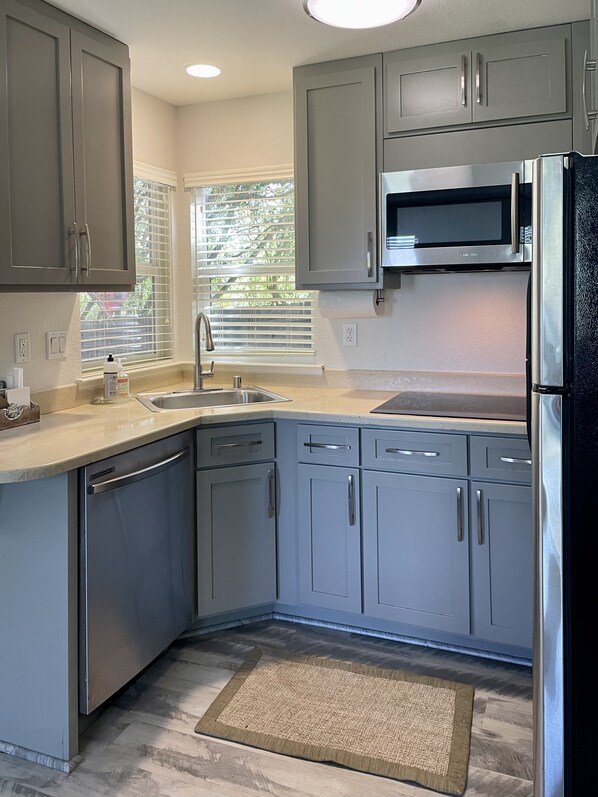 A modern kitchen with grey cabinets, stainless steel appliances, a window with blinds, and a wooden floor.