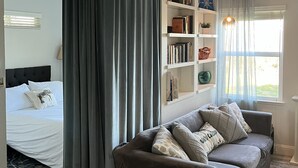 A room with a bed, a grey sofa, a bookshelf, and a window with curtains.