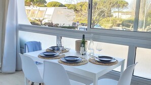 A dining table set with plates, glasses, and a bottle, surrounded by white chairs with wooden legs, in a room with large windows. - Renovated Boho Studio ☀ Pool | Beach | Queen-size bed (La Grande Motte)