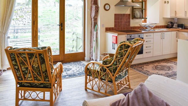 A cozy room with a wooden door leading to a garden, wicker chairs, a bed, a kitchen with white cabinets, and a clock on the wall.