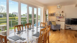 A spacious living area with a dining table, chairs, a sofa, and a TV. Large windows offer a view of the garden and neighboring houses. - An eco-friendly holiday bungalow near Abererch Beach on the Llyn Peninsula. (Pwllheli)
