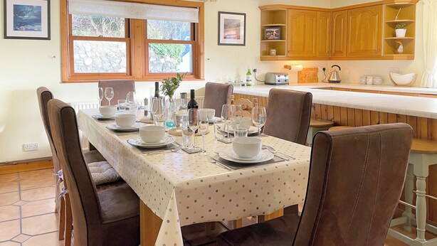A dining area with a table set for four, chairs, a kitchen in the background, and a window with a view.