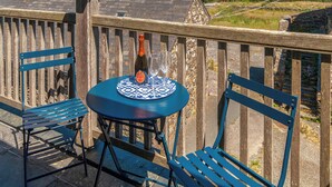 Une table avec une bouteille et des verres posés sur une nappe bleue, deux chaises bleues et une vue sur un paysage verdoyant.